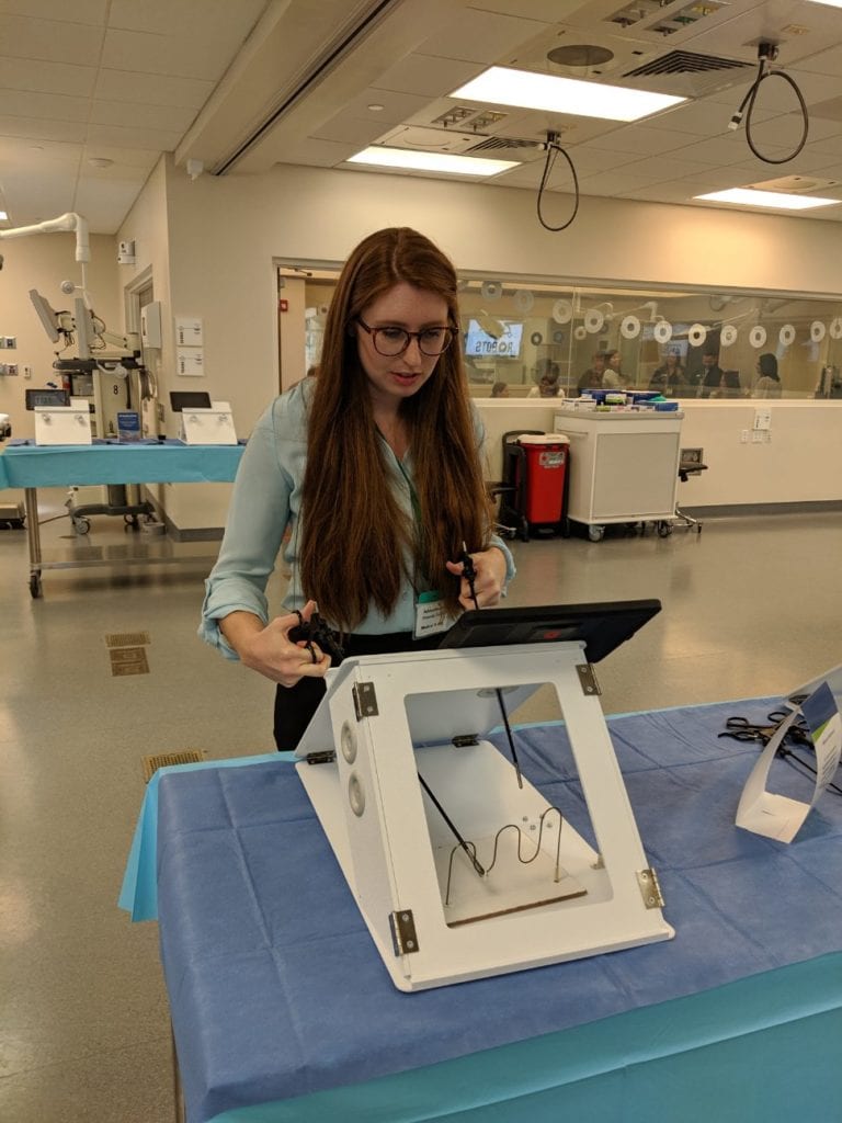 Halldale Group Industry Reporter Amanda Towner practices her dexterity skills, moving a ring through a looping metal wire of a puzzle with laparoscopic instruments. Image credit: Author.